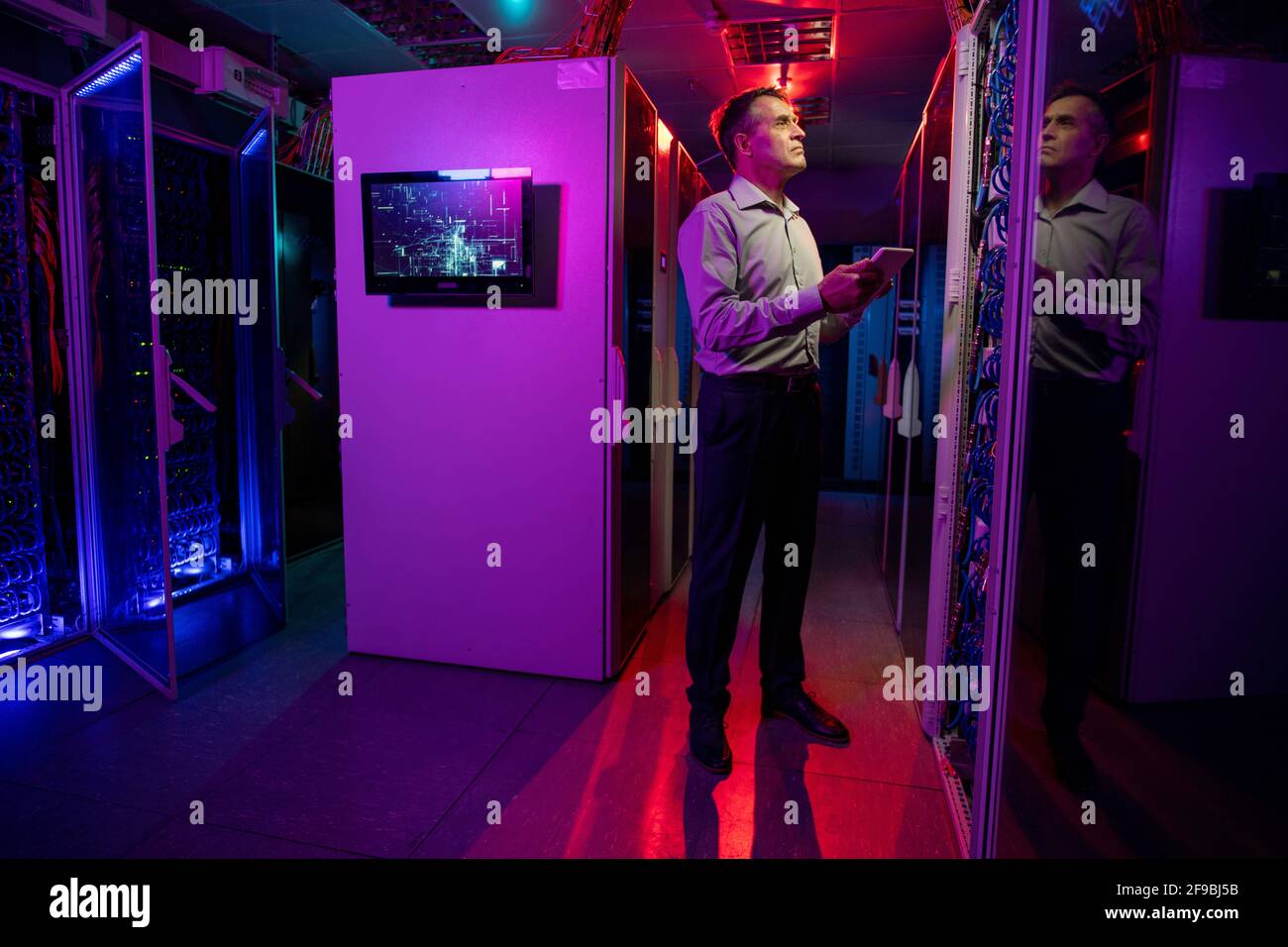 Ingénieur INFORMATIQUE masculin concentré debout dans une pièce au néon foncé pleine des armoires de serveurs et de l'utilisation d'une tablette tout en contrôlant les systèmes réseau Banque D'Images