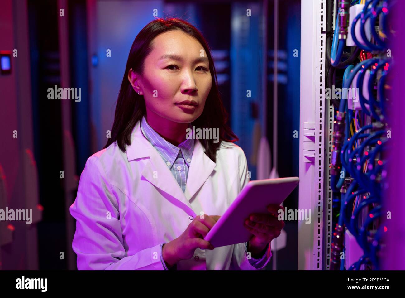 Portrait de contenu en blouse de laboratoire, jeune ingénieur INFORMATIQUE asiatique utilisation de la tablette lors du dépannage d'un problème de serveur sous lumière néon Banque D'Images