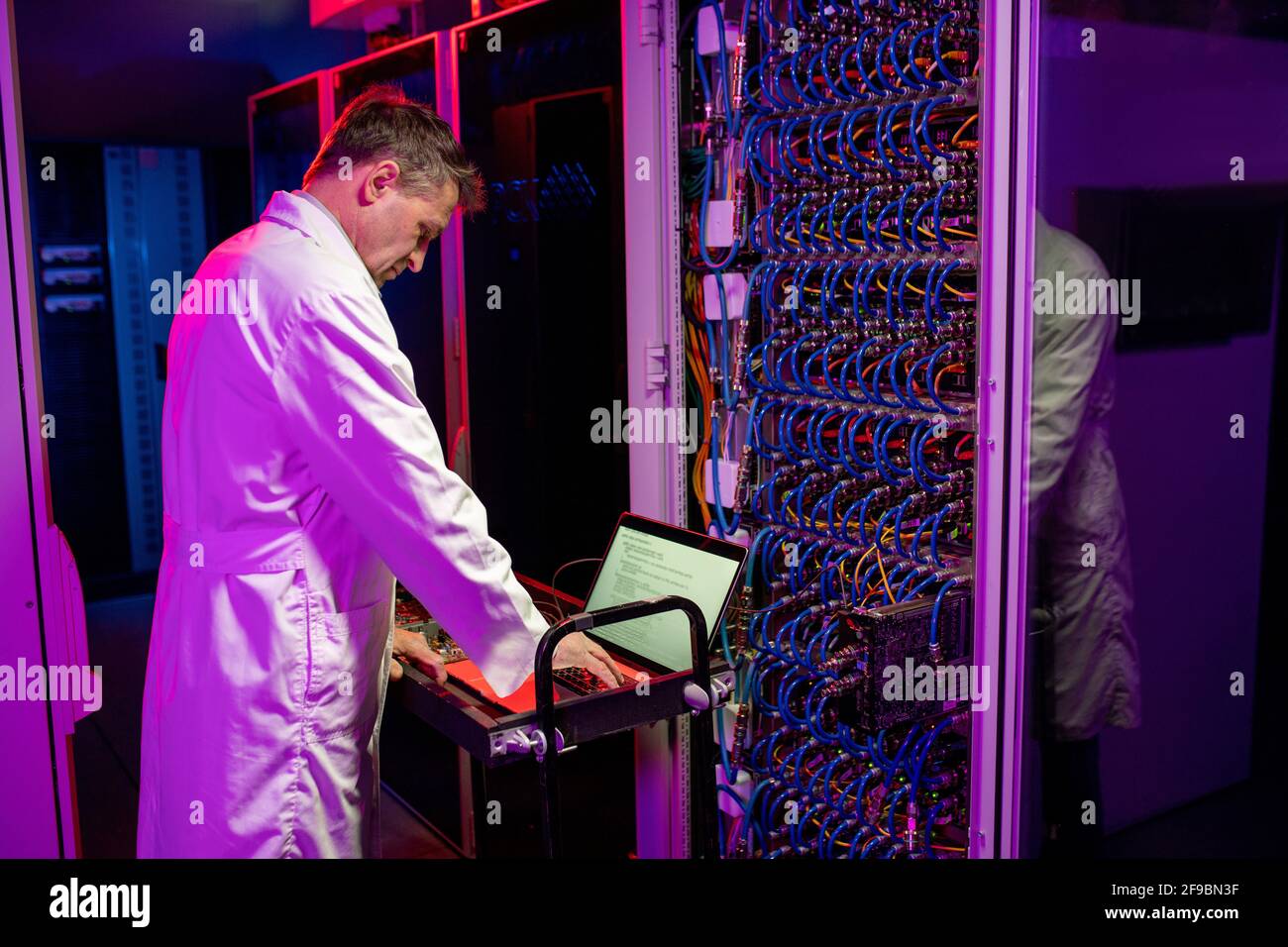 Ingénieur réseau mature concentré en couche blanche debout sur le chariot avec ordinateur portable et serveur lame de configuration dans une pièce éclairée au néon Banque D'Images