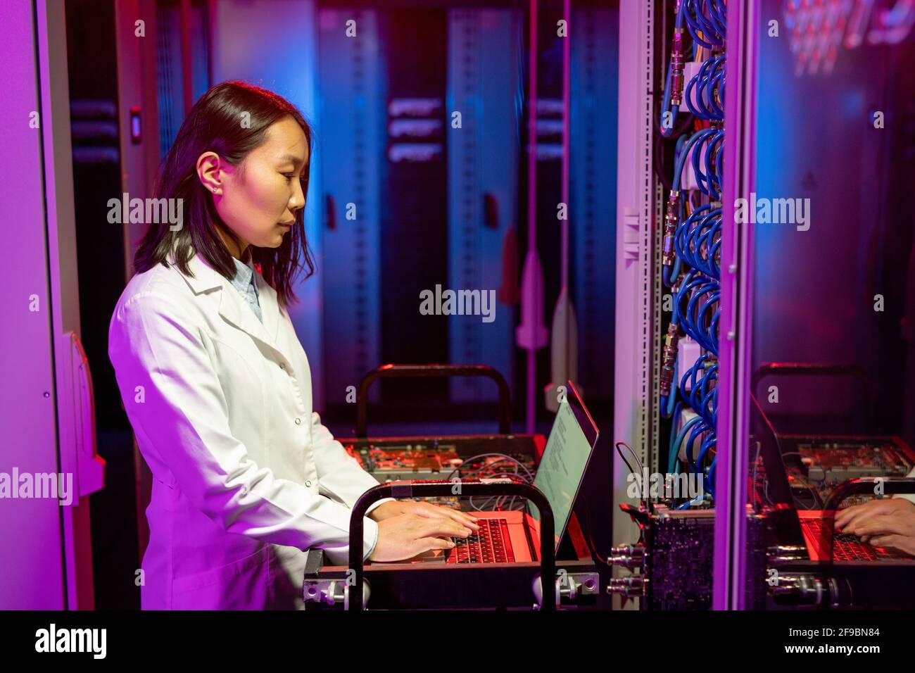 Jeune spécialiste INFORMATIQUE asiatique sérieux en blouse blanche chariot et utilisation de l'ordinateur portable lors de l'exécution des tests de diagnostic Banque D'Images