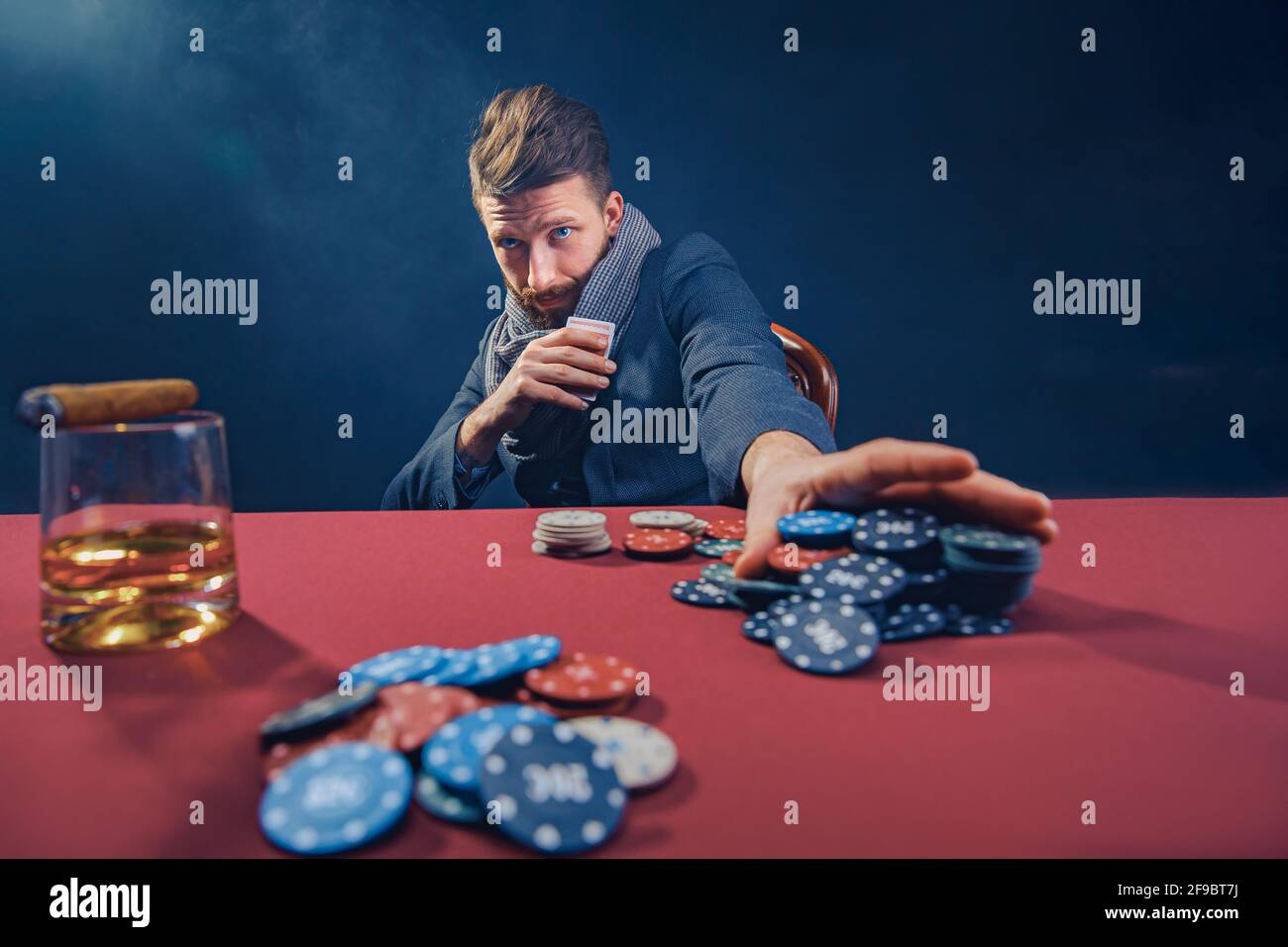 Elégant homme barbu en costume et foulard jouant dans le casino sombre, fume cigare, boisson whisky Banque D'Images