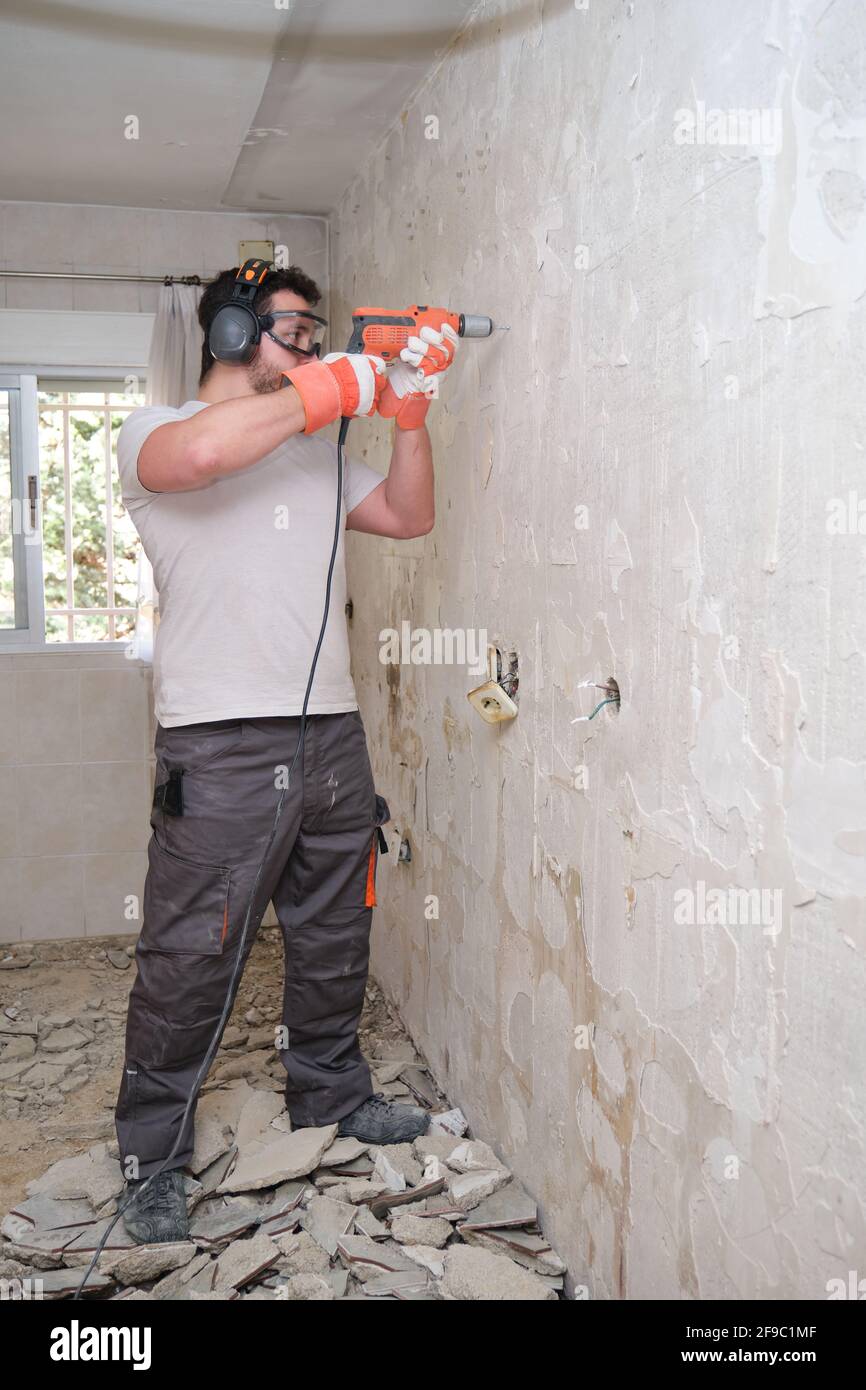 Constructeur forant des trous dans un mur avec une perceuse électrique sur le chantier de construction. Travailleur de la construction portant des gants de sécurité, des lunettes et des couvre-oreilles. Banque D'Images