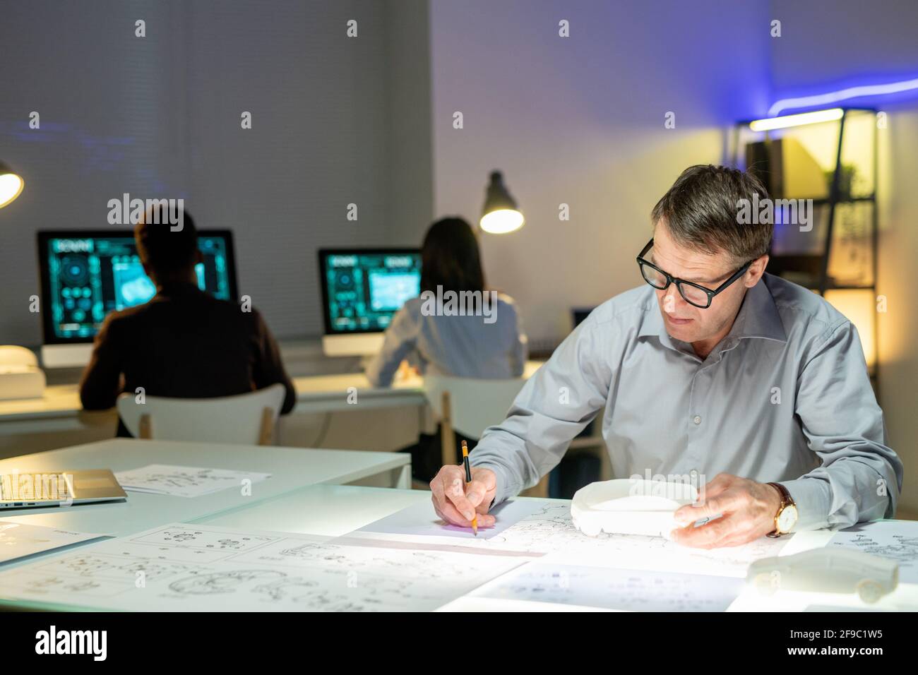 Concepteur automobile mature concentré en lunettes assises avec le modèle de voiture à la table et modification des mises en page dans le bureau Banque D'Images