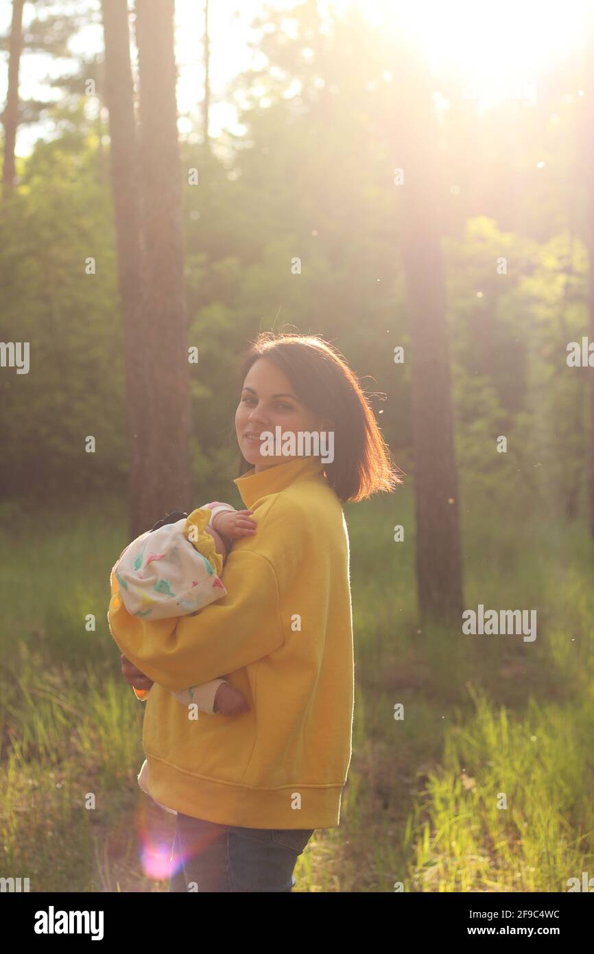 Une belle jeune mère avec des cheveux droits et dans un le sweat-shirt jaune tient un petit enfant dans ses bras la forêt dans l'éblouissement du soleil du soir Banque D'Images