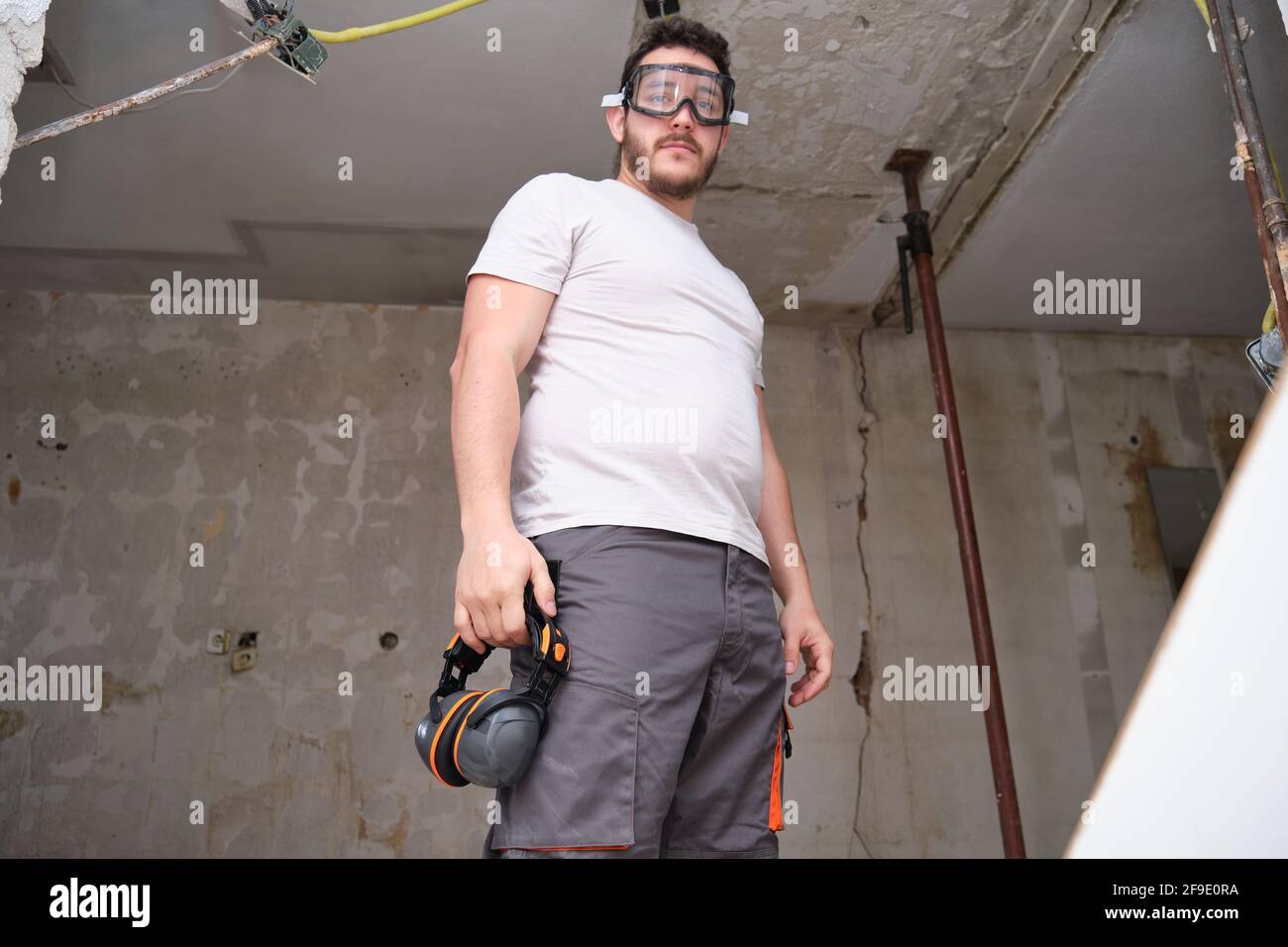Un jeune constructeur porte des lunettes de sécurité sur un chantier de construction. Protections auditives suspendues au pantalon. Banque D'Images