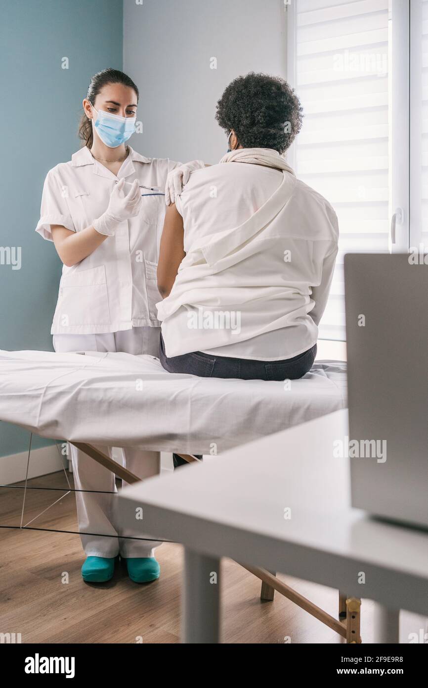 Femme médecin spécialiste en uniforme de protection, gants en latex et masque facial vaccinant la patiente africaine américaine mature en clinique pendant le coronav Banque D'Images