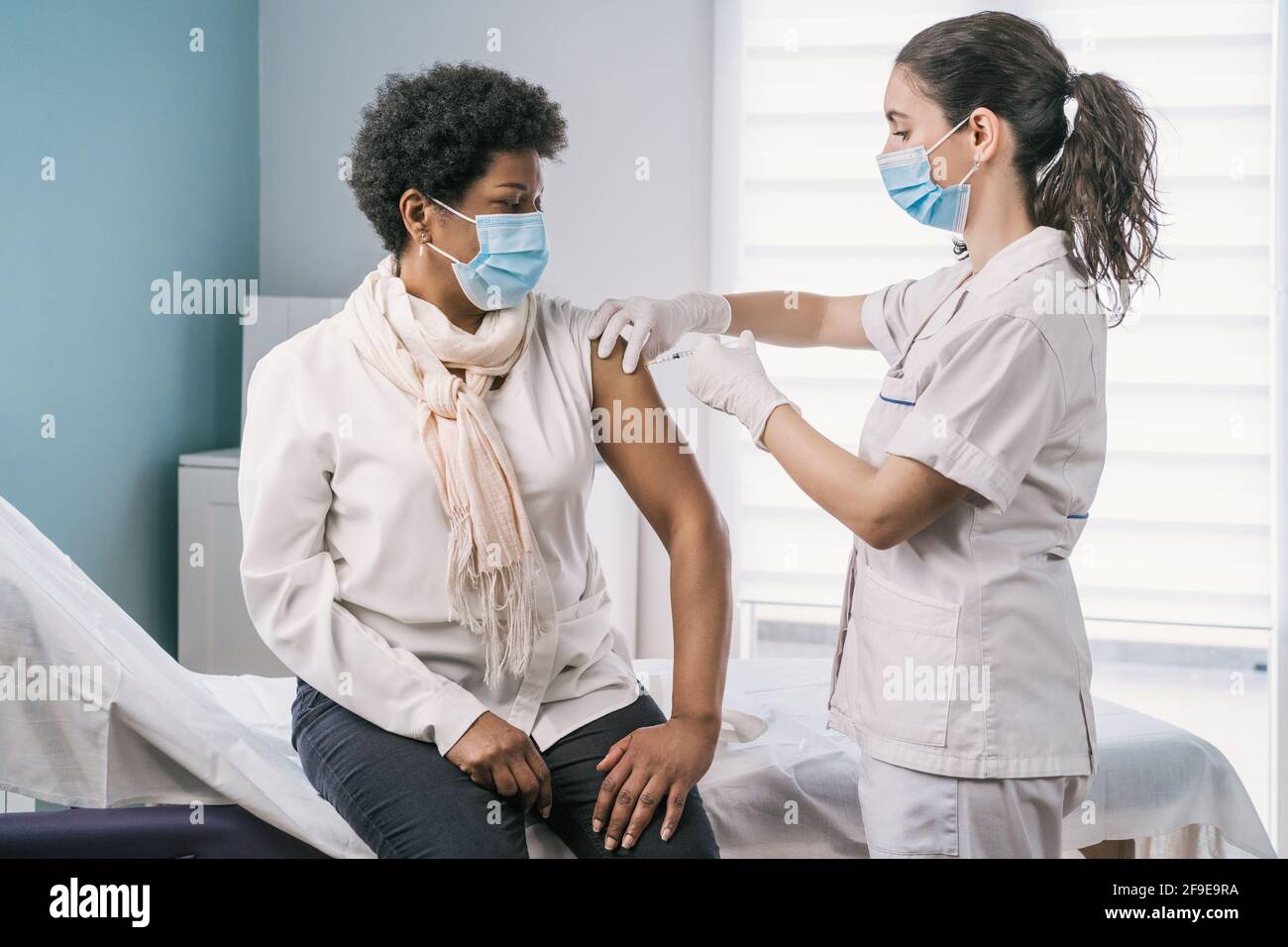 Femme médecin spécialiste en uniforme de protection, gants en latex et masque facial vaccinant la patiente africaine américaine mature en clinique pendant le coronav Banque D'Images