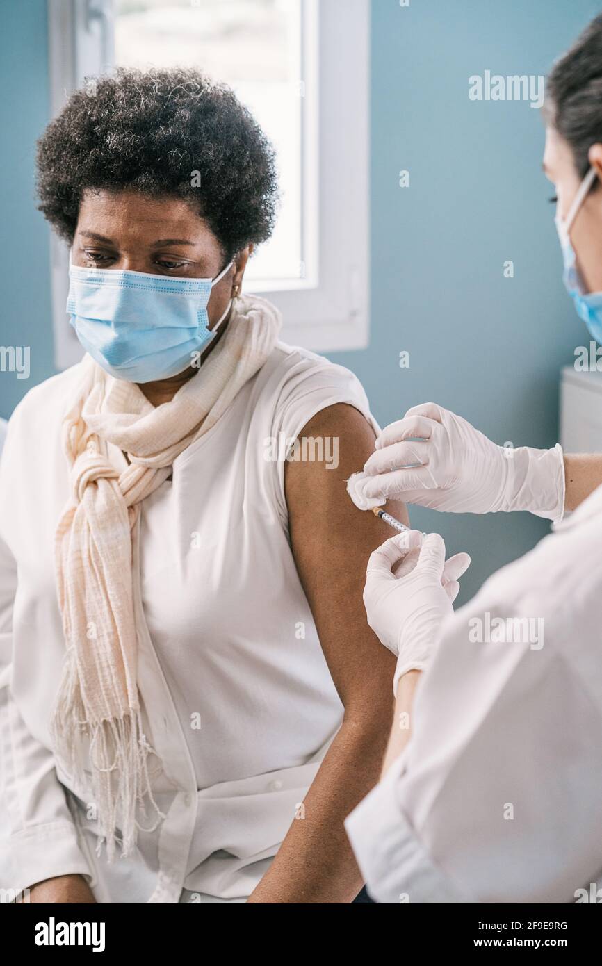 Femme médecin spécialiste en uniforme de protection, gants en latex et masque facial vaccinant la patiente africaine américaine mature en clinique pendant le coronav Banque D'Images