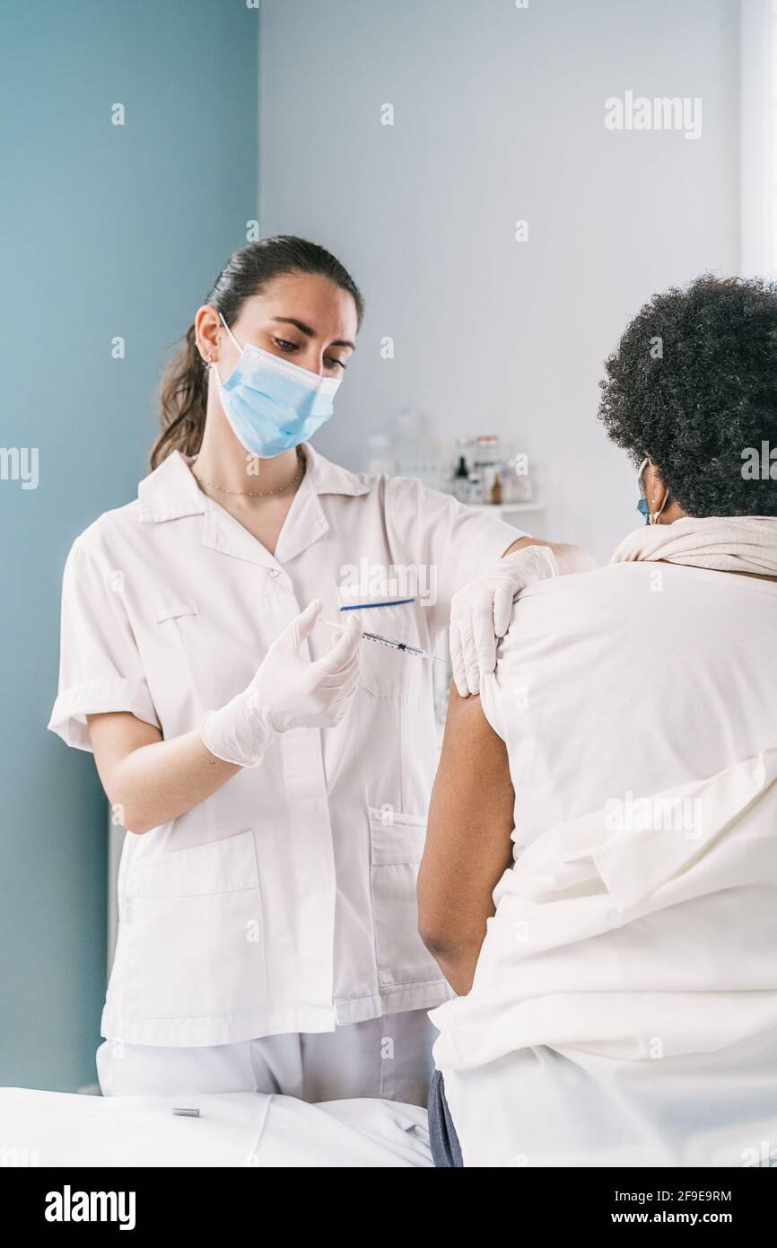 Femme médecin spécialiste en uniforme de protection, gants en latex et masque facial vaccinant la patiente africaine américaine mature en clinique pendant le coronav Banque D'Images