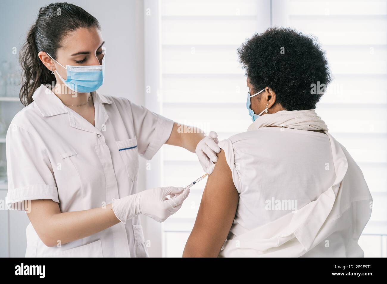Femme médecin spécialiste en uniforme de protection, gants en latex et masque facial vaccinant la patiente africaine américaine mature en clinique pendant le coronav Banque D'Images