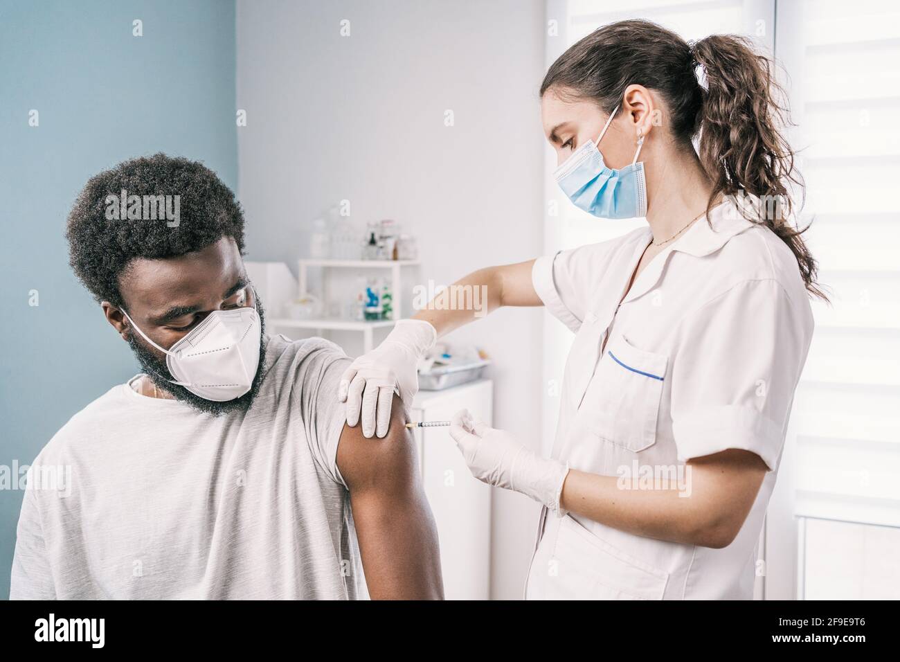 Femme médecin spécialiste en uniforme de protection, gants en latex et masque facial vaccinant l'homme afro-américain en clinique pendant l'outb du coronavirus Banque D'Images