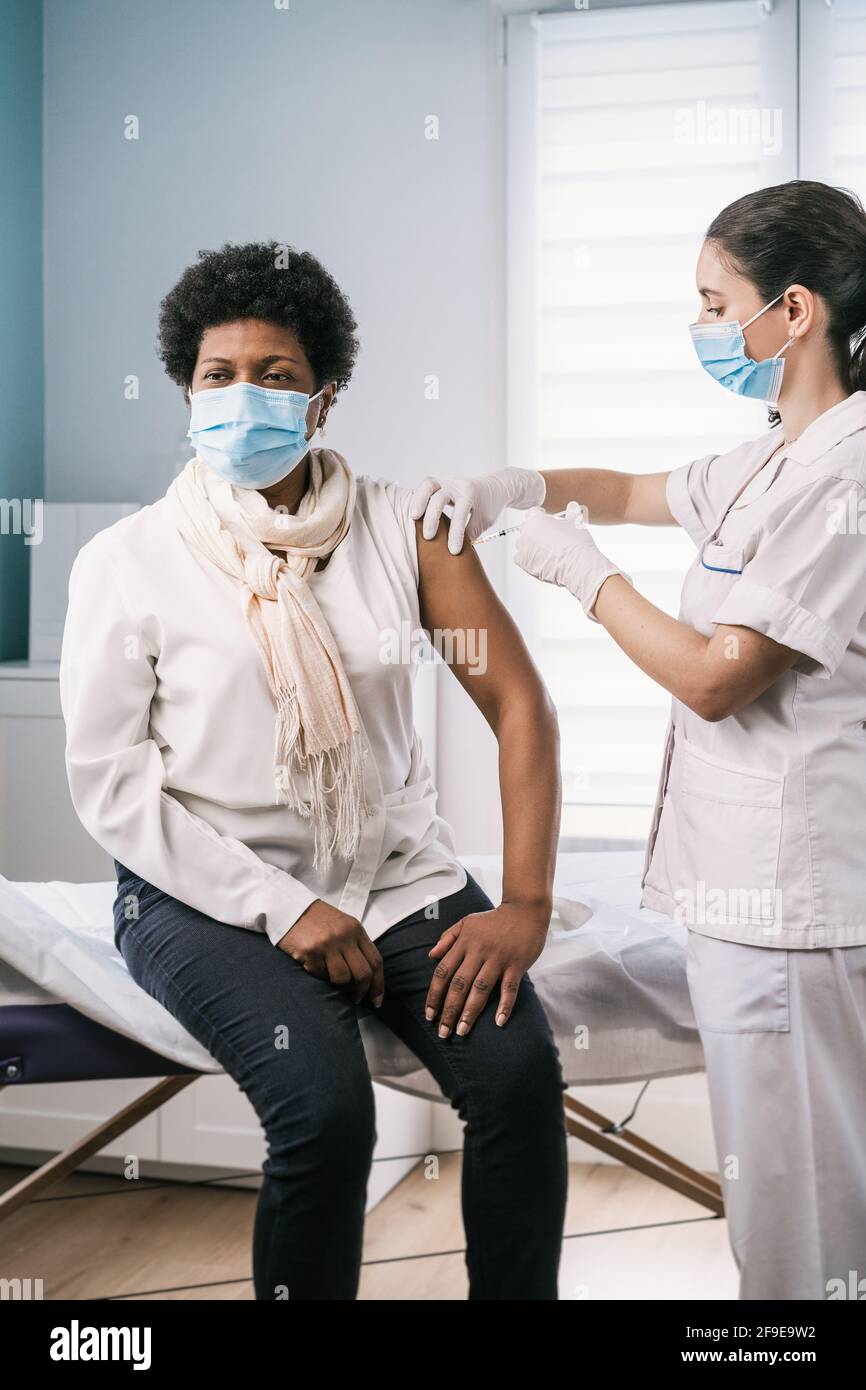 Femme médecin spécialiste en uniforme de protection, gants en latex et masque facial vaccinant la patiente africaine américaine mature en clinique pendant le coronav Banque D'Images