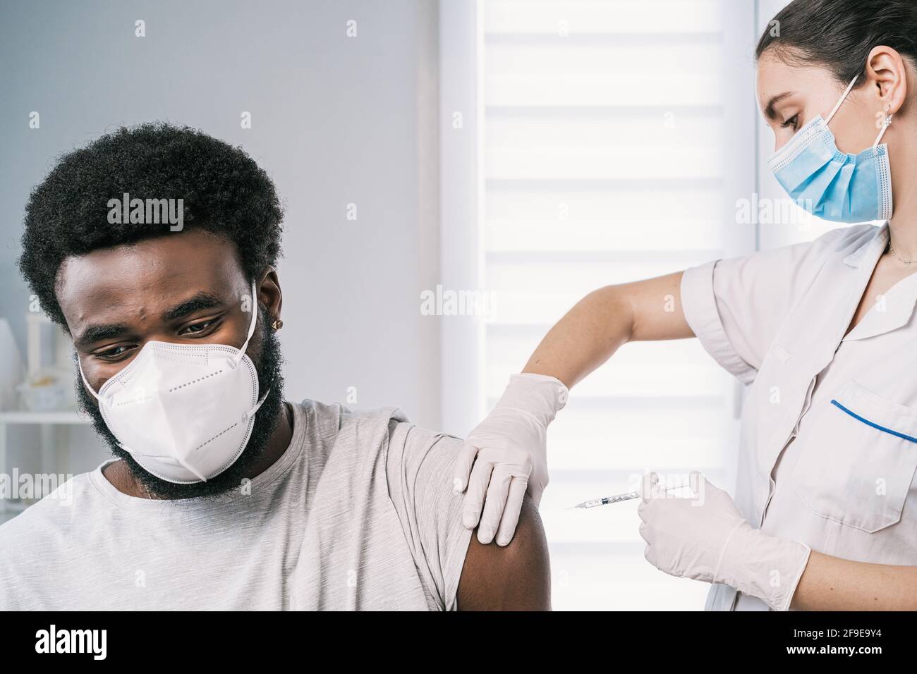 Femme médecin spécialiste en uniforme de protection, gants en latex et masque facial vaccinant l'homme afro-américain en clinique pendant l'outb du coronavirus Banque D'Images