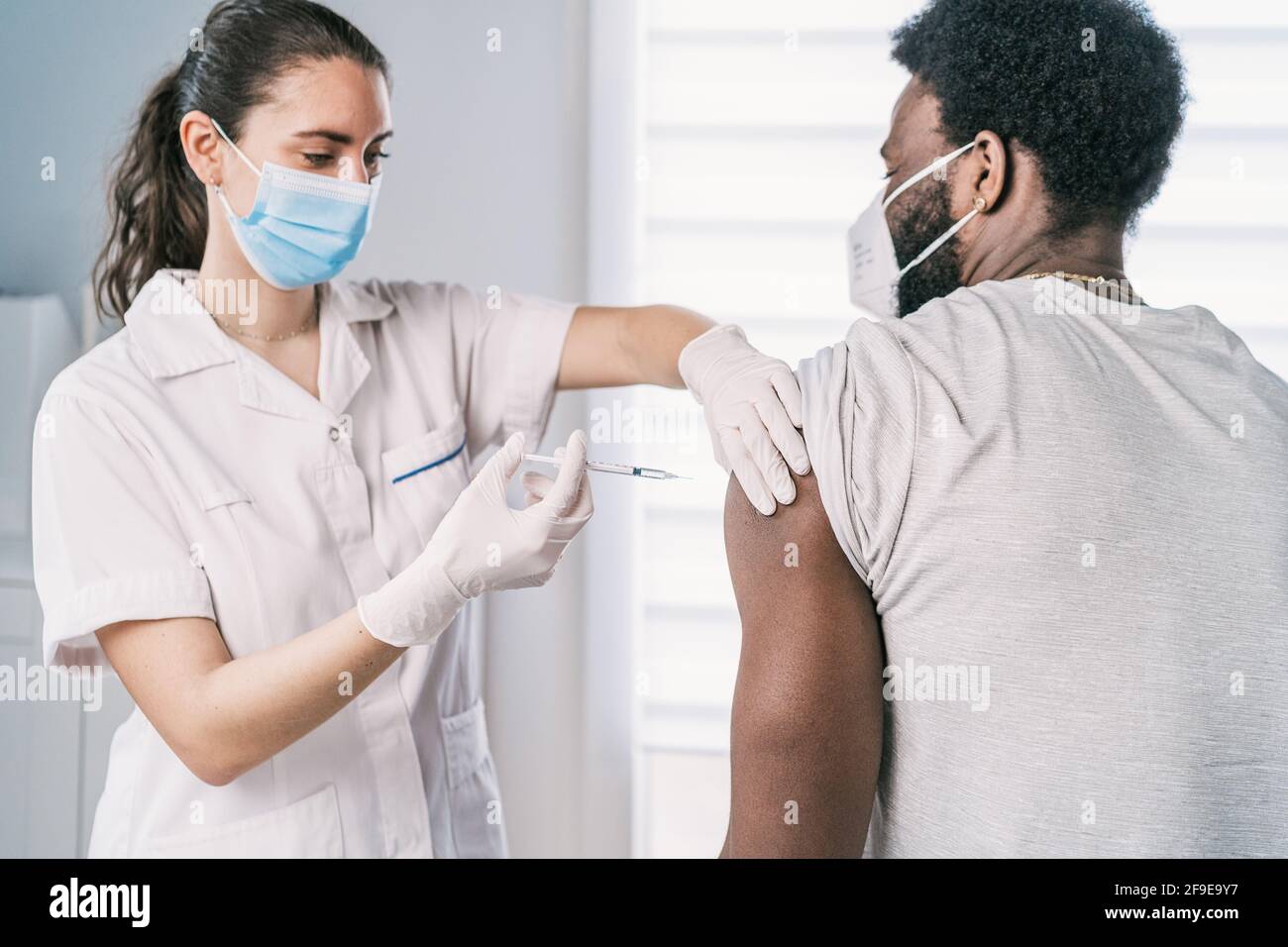 Femme médecin spécialiste en uniforme de protection, gants en latex et masque facial vaccinant l'homme afro-américain en clinique pendant l'outb du coronavirus Banque D'Images