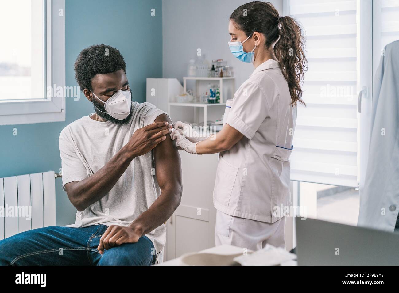 Femme médecin spécialiste en uniforme de protection, gants en latex et masque facial vaccinant l'homme afro-américain en clinique pendant l'outb du coronavirus Banque D'Images