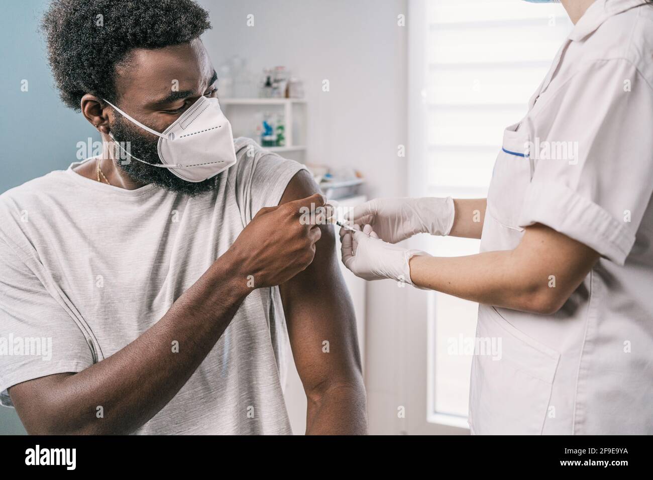 Spécialiste médical féminin en uniforme de protection, gants en latex et masque facial, non reconnaissable, en culture, qui vaccine un homme afro-américain en clinique Banque D'Images