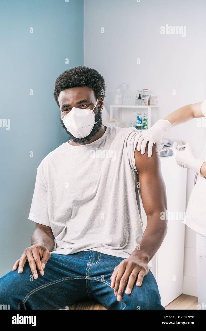 Spécialiste médical féminin en uniforme de protection, gants en latex et masque facial, non reconnaissable, en culture, qui vaccine un homme afro-américain en clinique Banque D'Images