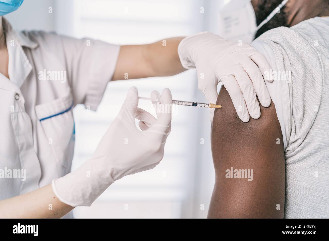 Spécialiste médical féminin en uniforme de protection, gants en latex et masque facial, non reconnaissable, en culture, qui vaccine un homme afro-américain en clinique Banque D'Images