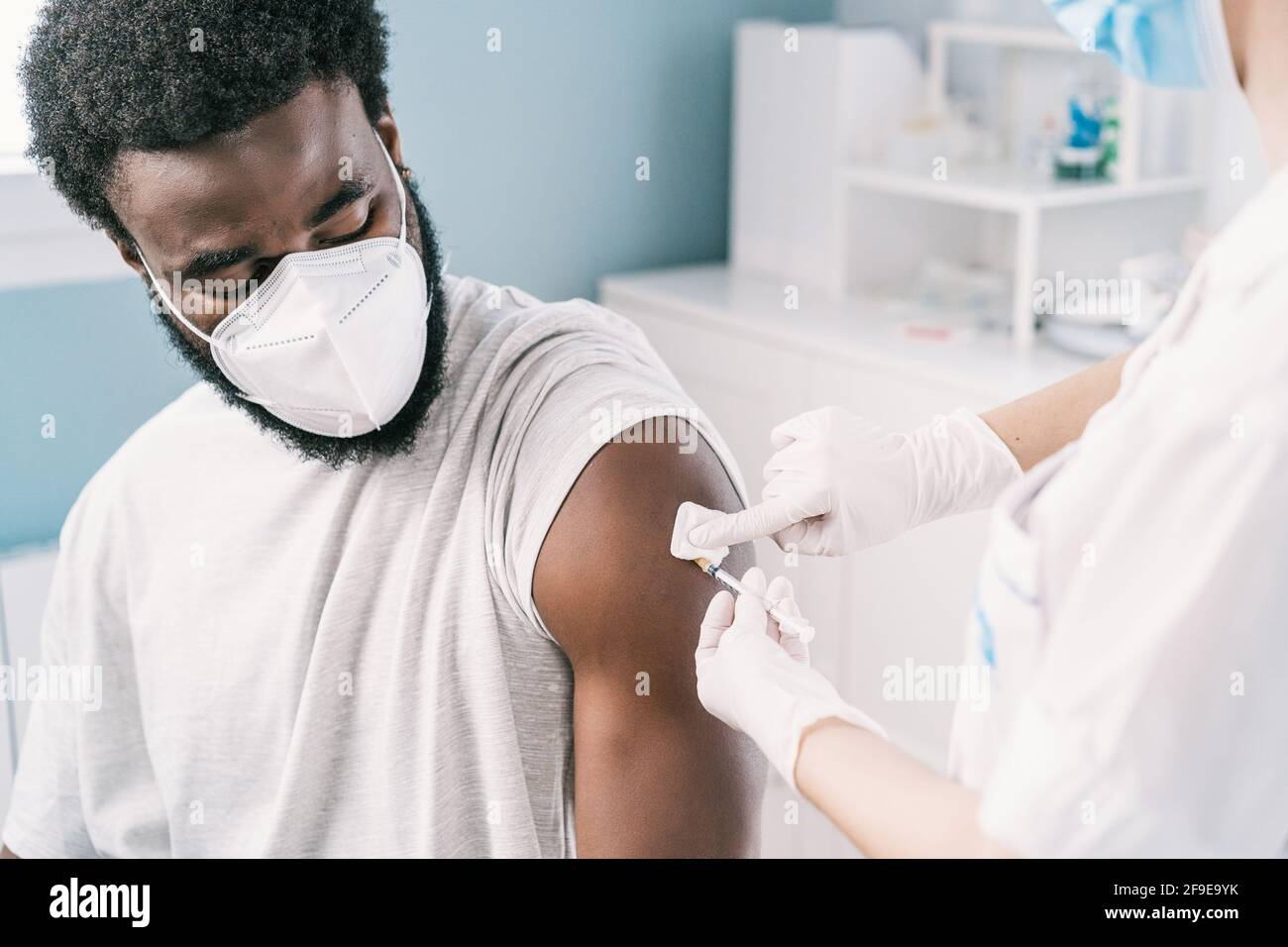 Spécialiste médical féminin en uniforme de protection, gants en latex et masque facial, non reconnaissable, en culture, qui vaccine un homme afro-américain en clinique Banque D'Images