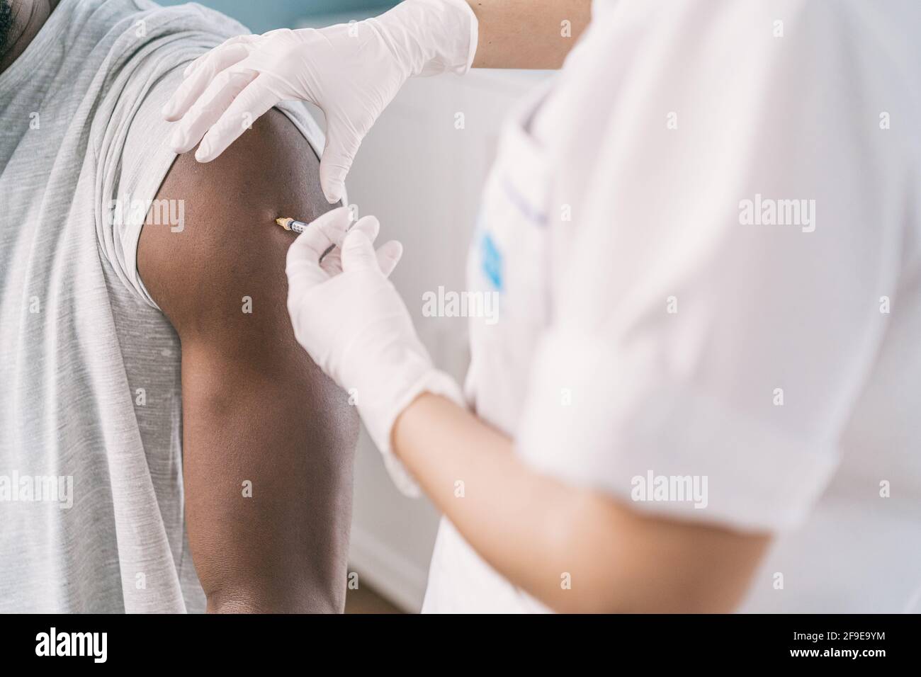 Spécialiste médical féminin en uniforme de protection, gants en latex et masque facial, non reconnaissable, en culture, qui vaccine un homme afro-américain en clinique Banque D'Images