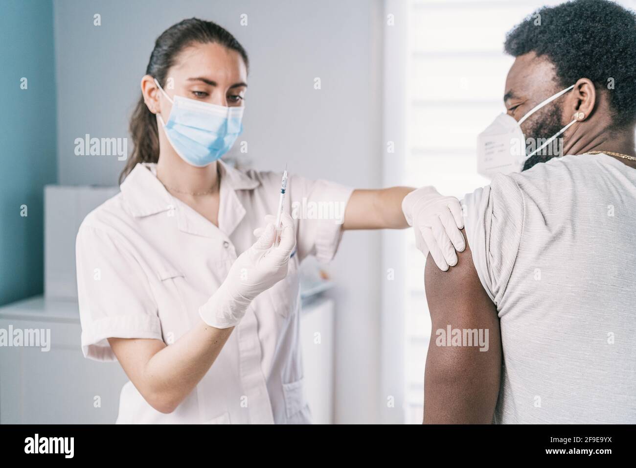 Femme médecin spécialiste en uniforme de protection, gants en latex et masque facial vaccinant l'homme afro-américain en clinique pendant l'outb du coronavirus Banque D'Images