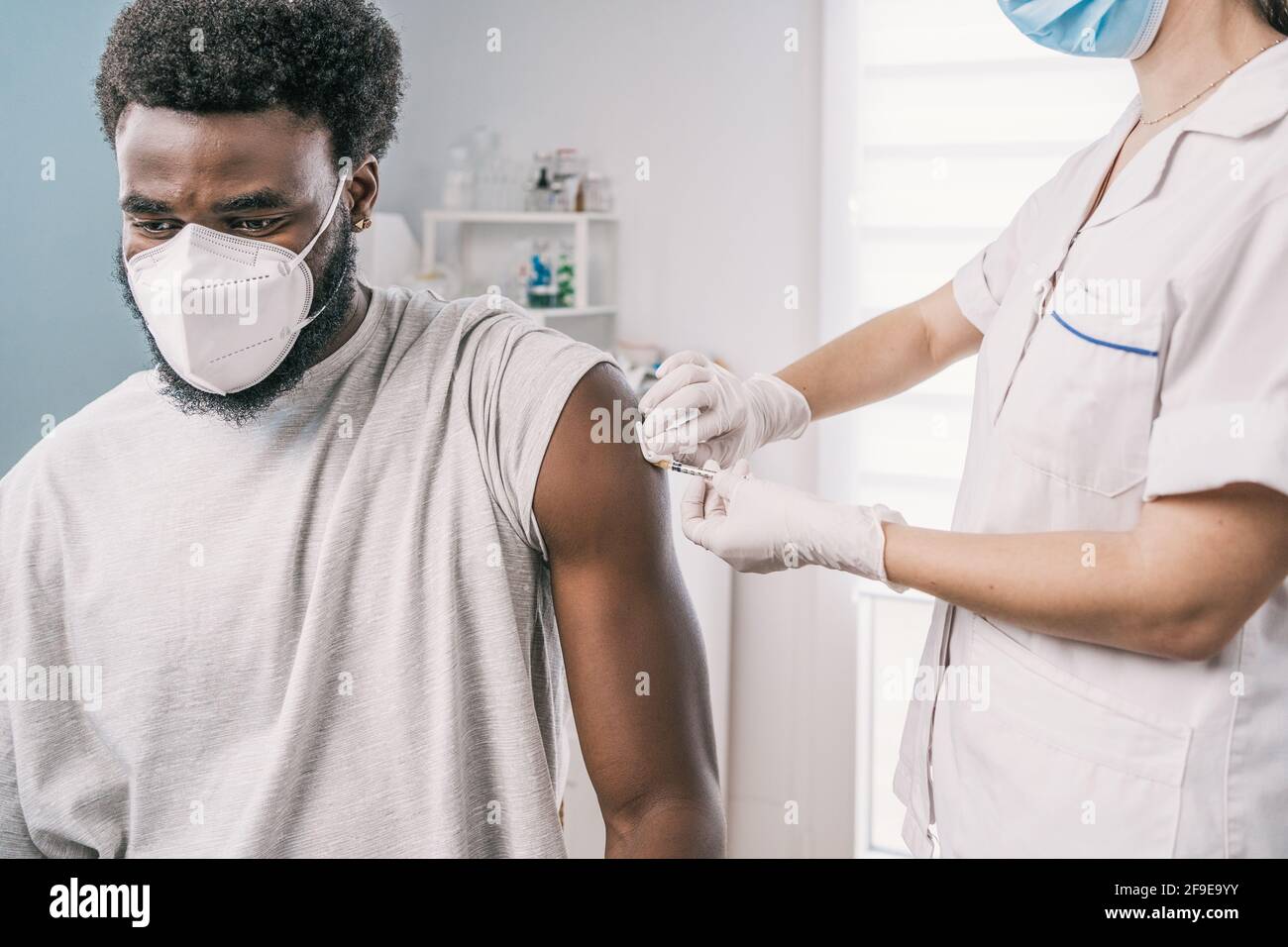 Spécialiste médical féminin en uniforme de protection, gants en latex et masque facial, non reconnaissable, en culture, qui vaccine un homme afro-américain en clinique Banque D'Images