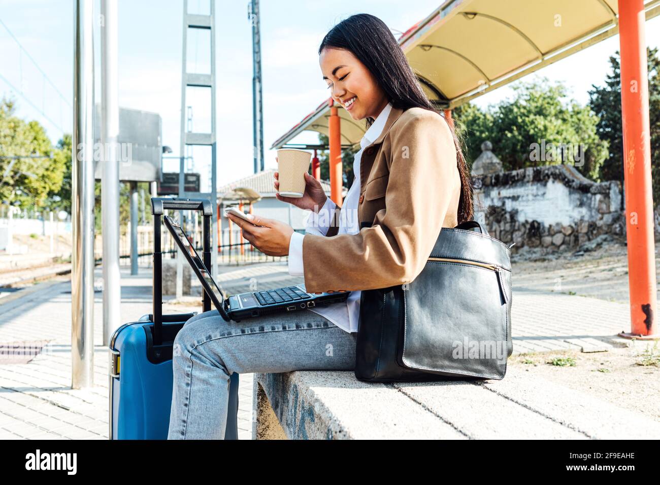 Vue latérale d'une femme asiatique pigiste assise sur un banc avec ordinateur portable et café à emporter et navigation sur smartphone pendant que vous attendez pour le train à chemin de fer Banque D'Images