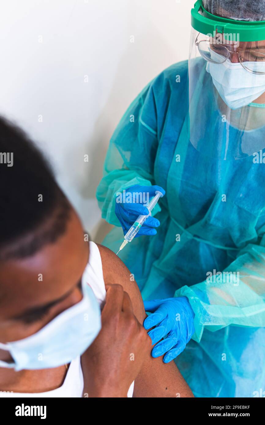 Spécialiste médical féminin en uniforme de protection, gants en latex et masque facial, non reconnaissable, en culture, qui vaccine un homme afro-américain en clinique Banque D'Images