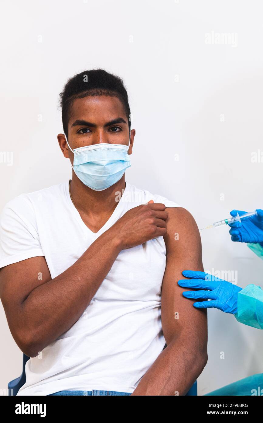 Spécialiste médical féminin en uniforme de protection, gants en latex et masque facial, non reconnaissable, en culture, qui vaccine un homme afro-américain en clinique Banque D'Images