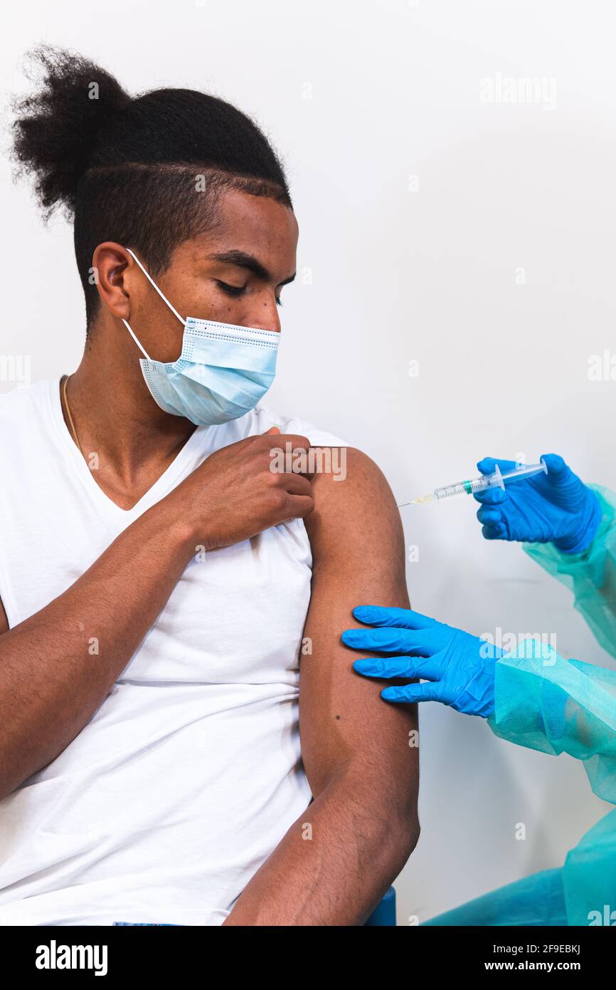 Spécialiste médical féminin en uniforme de protection, gants en latex et masque facial, non reconnaissable, en culture, qui vaccine un homme afro-américain en clinique Banque D'Images