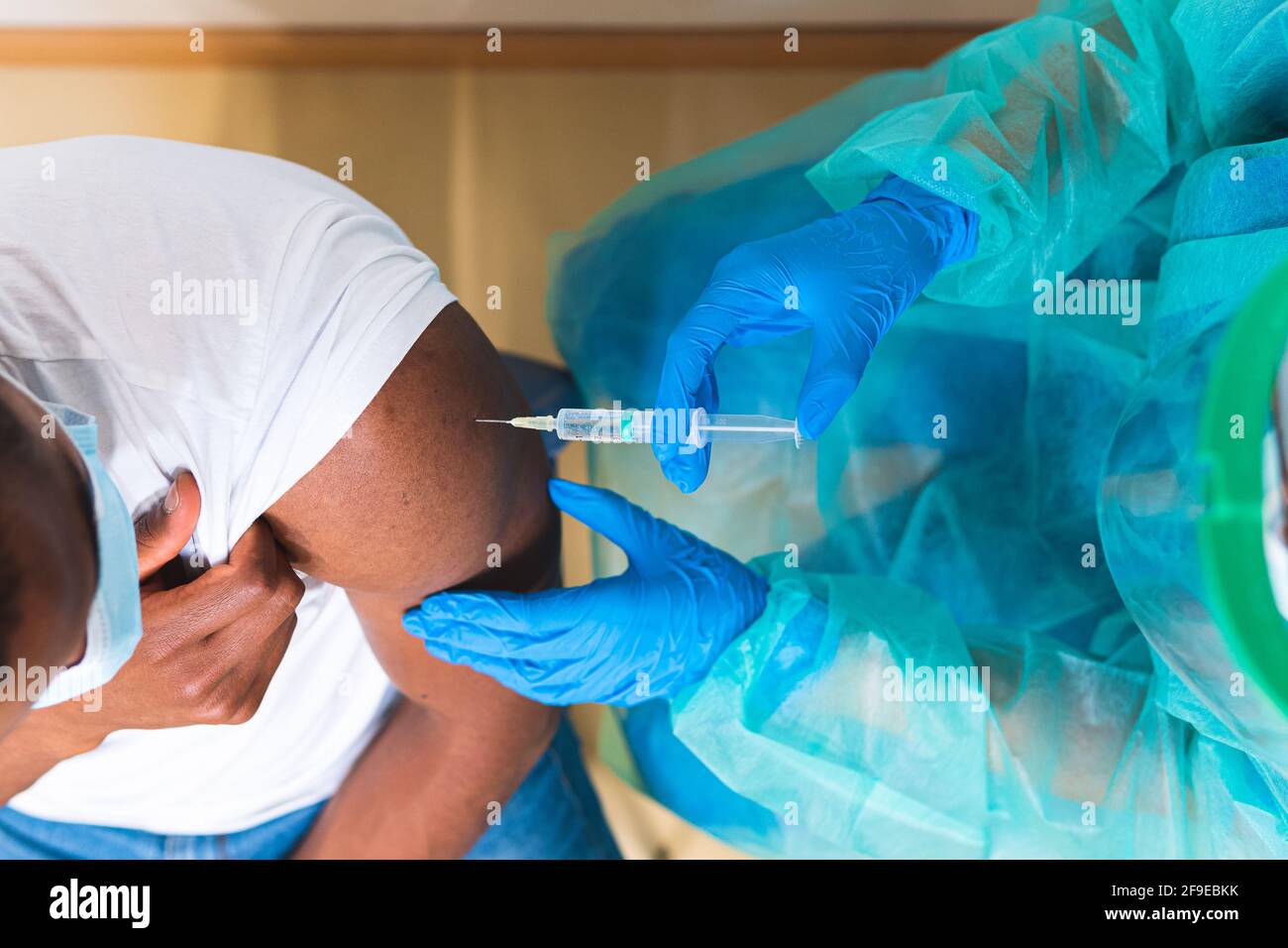 Spécialiste médical féminin en uniforme de protection, gants en latex et masque facial, non reconnaissable, en culture, qui vaccine un homme afro-américain en clinique Banque D'Images