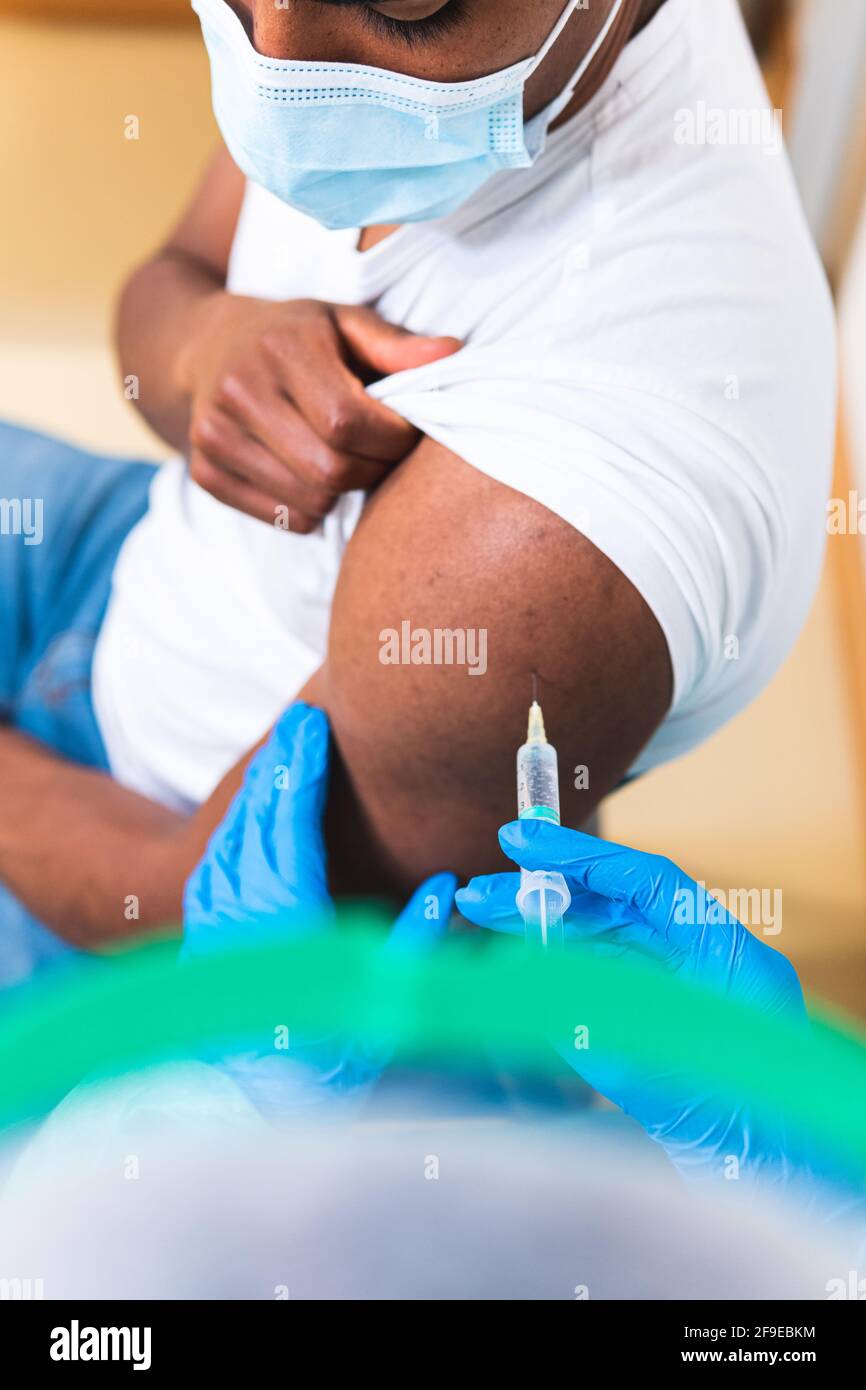 Spécialiste médical féminin en uniforme de protection, gants en latex et masque facial, non reconnaissable, en culture, qui vaccine un homme afro-américain en clinique Banque D'Images