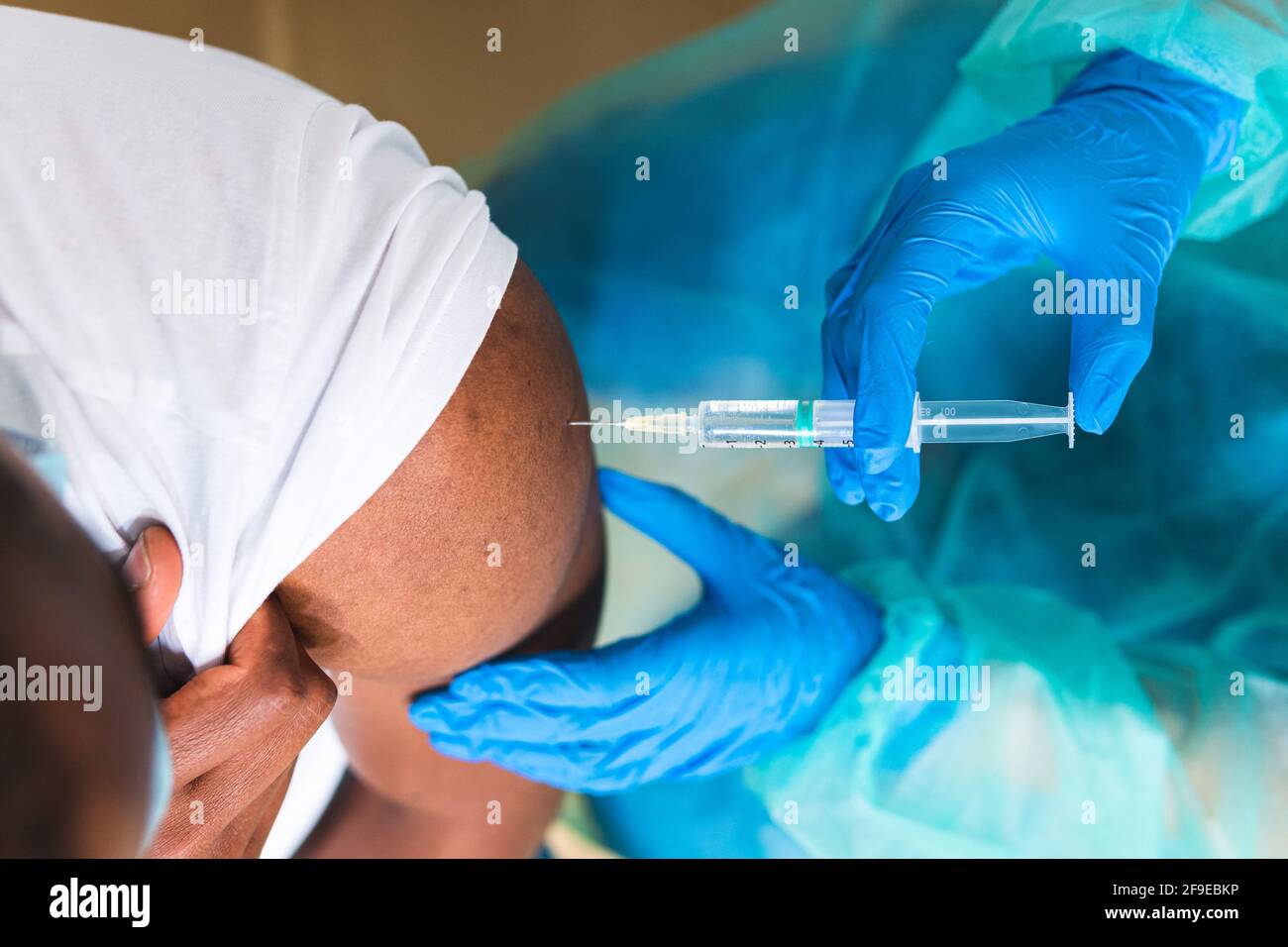 Spécialiste médical féminin en uniforme de protection, gants en latex et masque facial, non reconnaissable, en culture, qui vaccine un homme afro-américain en clinique Banque D'Images