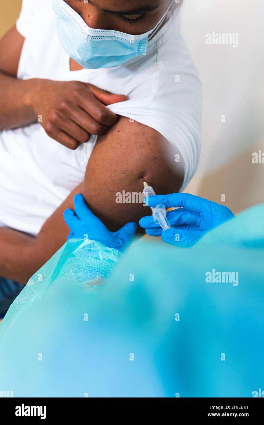 Spécialiste médical féminin en uniforme de protection, gants en latex et masque facial, non reconnaissable, en culture, qui vaccine un homme afro-américain en clinique Banque D'Images