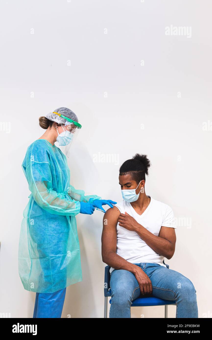 Femme médecin spécialiste en uniforme de protection, gants en latex et masque facial vaccinant l'homme afro-américain en clinique pendant l'outb du coronavirus Banque D'Images