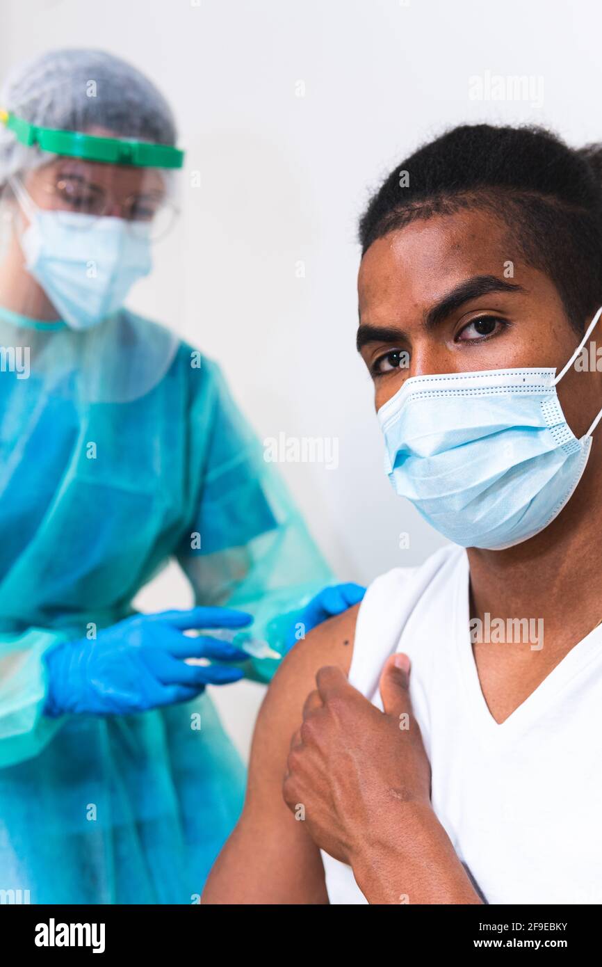 Femme médecin spécialiste en uniforme de protection, gants en latex et masque facial vaccinant l'homme afro-américain en clinique pendant l'outb du coronavirus Banque D'Images