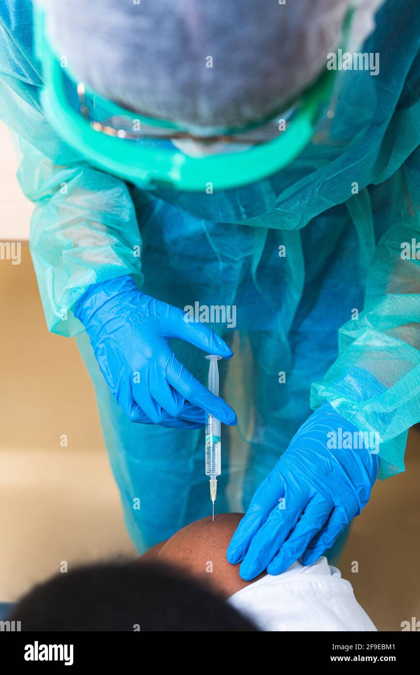Spécialiste médical féminin en uniforme de protection, gants en latex et masque facial, non reconnaissable, en culture, qui vaccine un homme afro-américain en clinique Banque D'Images