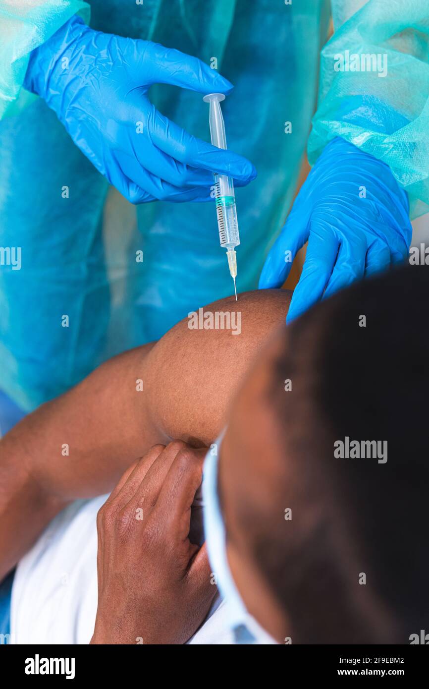 Spécialiste médical féminin en uniforme de protection, gants en latex et masque facial, non reconnaissable, en culture, qui vaccine un homme afro-américain en clinique Banque D'Images
