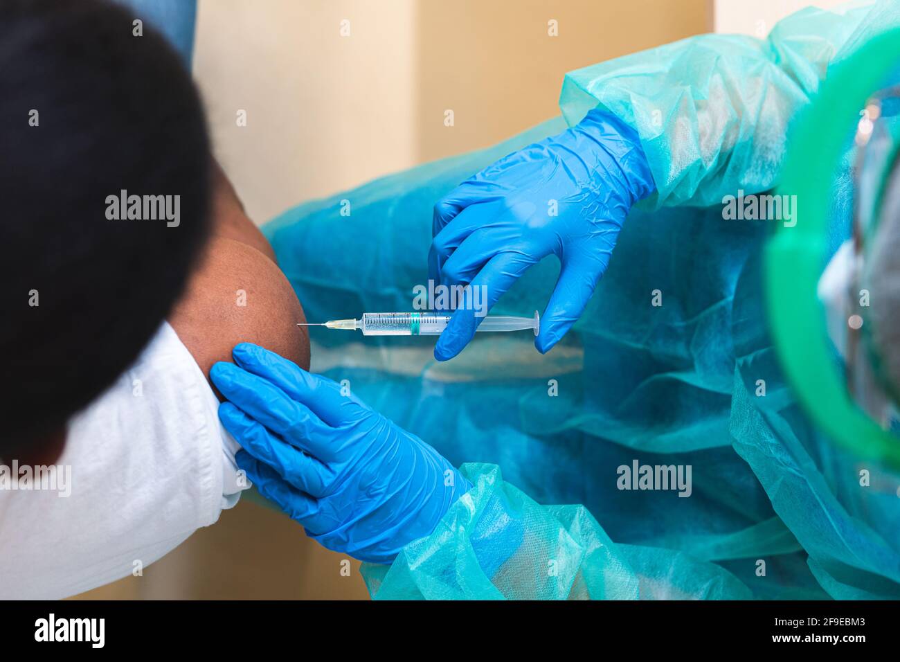 Spécialiste médical féminin en uniforme de protection, gants en latex et masque facial, non reconnaissable, en culture, qui vaccine un homme afro-américain en clinique Banque D'Images