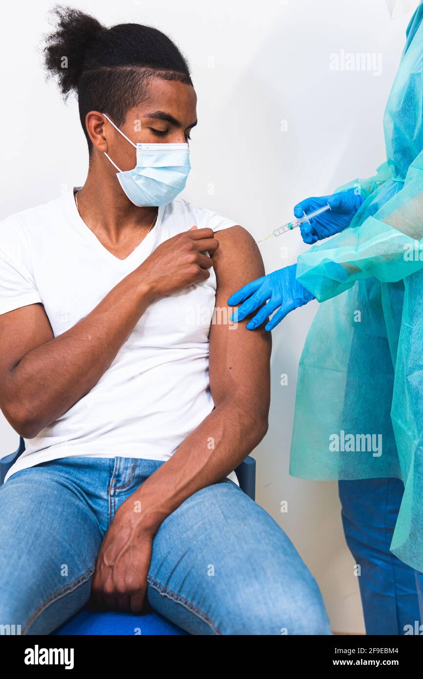 Spécialiste médical féminin en uniforme de protection, gants en latex et masque facial, non reconnaissable, en culture, qui vaccine un homme afro-américain en clinique Banque D'Images