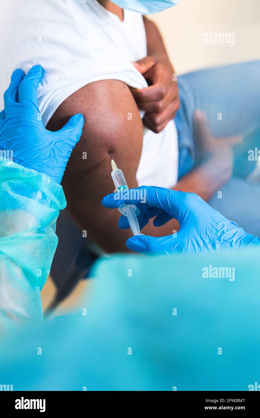 Spécialiste médical féminin en uniforme de protection, gants en latex et masque facial, non reconnaissable, en culture, qui vaccine un homme afro-américain en clinique Banque D'Images