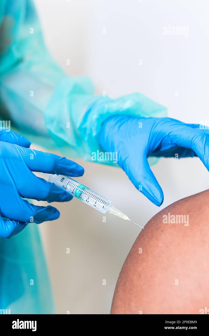 Spécialiste médical féminin en uniforme de protection, gants en latex et masque facial, non reconnaissable, en culture, qui vaccine un homme afro-américain en clinique Banque D'Images