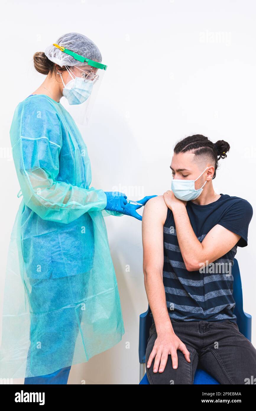 Spécialiste médical féminin en uniforme de protection, gants en latex et masque facial vaccinant un patient hispanique en clinique pendant une épidémie de coronavirus Banque D'Images
