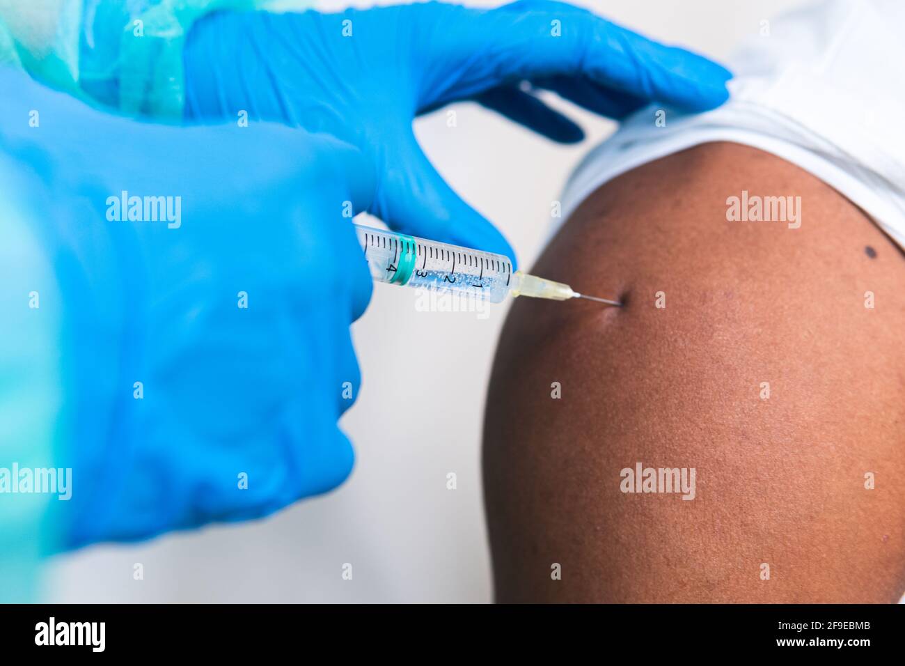 Spécialiste médical féminin en uniforme de protection, gants en latex et masque facial, non reconnaissable, en culture, qui vaccine un homme afro-américain en clinique Banque D'Images