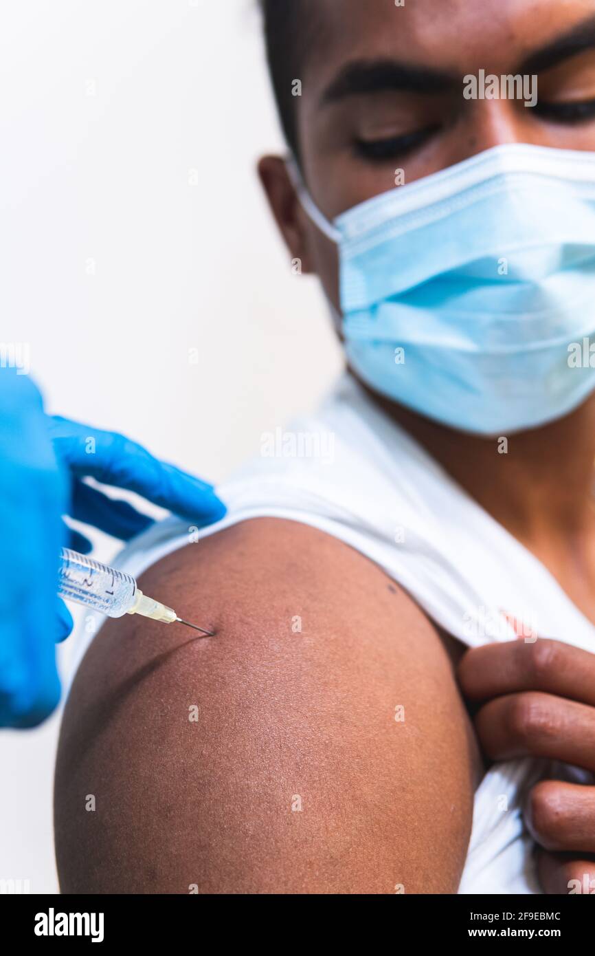 Spécialiste médical féminin en uniforme de protection, gants en latex et masque facial, non reconnaissable, en culture, qui vaccine un homme afro-américain en clinique Banque D'Images