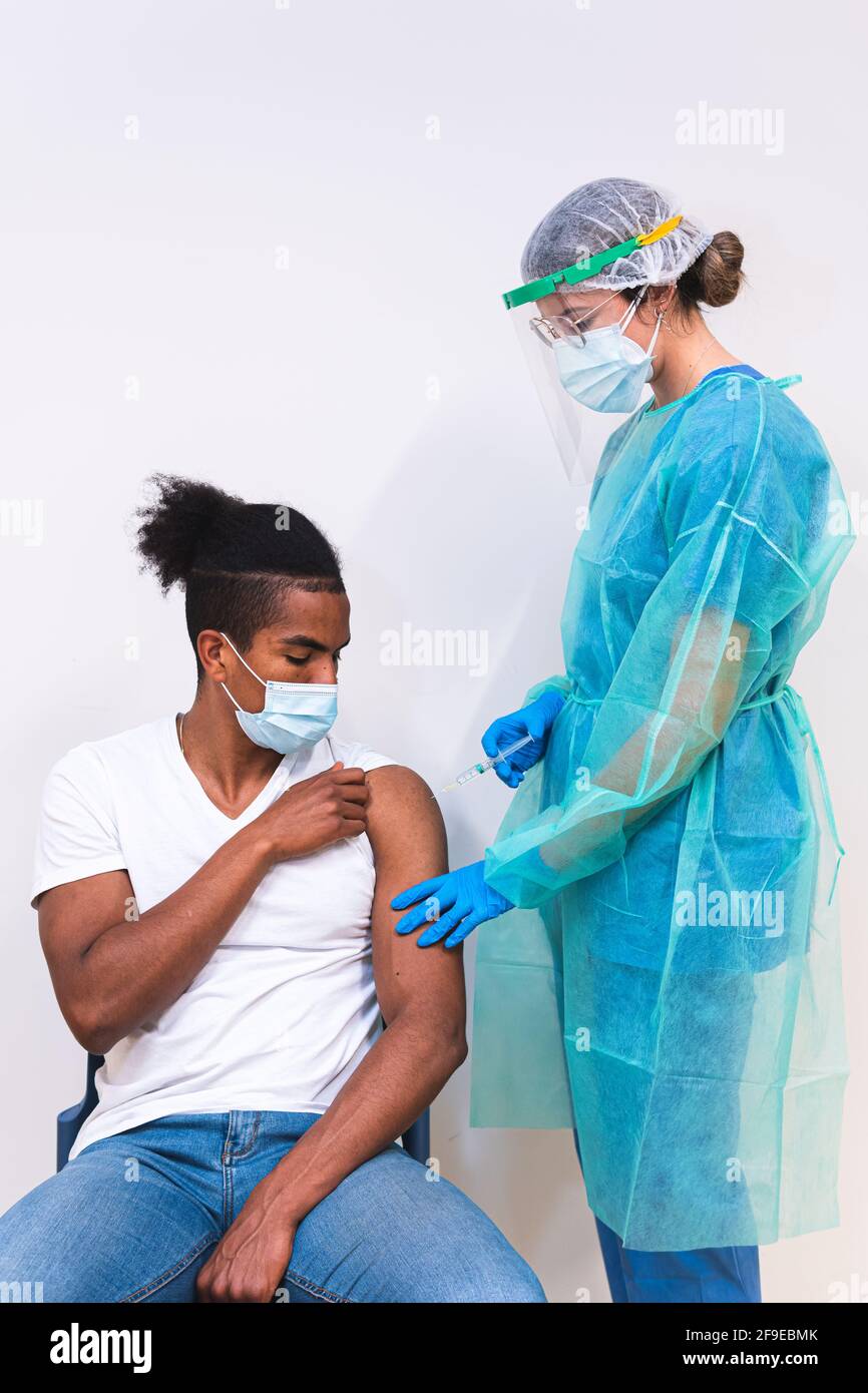Femme médecin spécialiste en uniforme de protection, gants en latex et masque facial vaccinant l'homme afro-américain en clinique pendant l'outb du coronavirus Banque D'Images