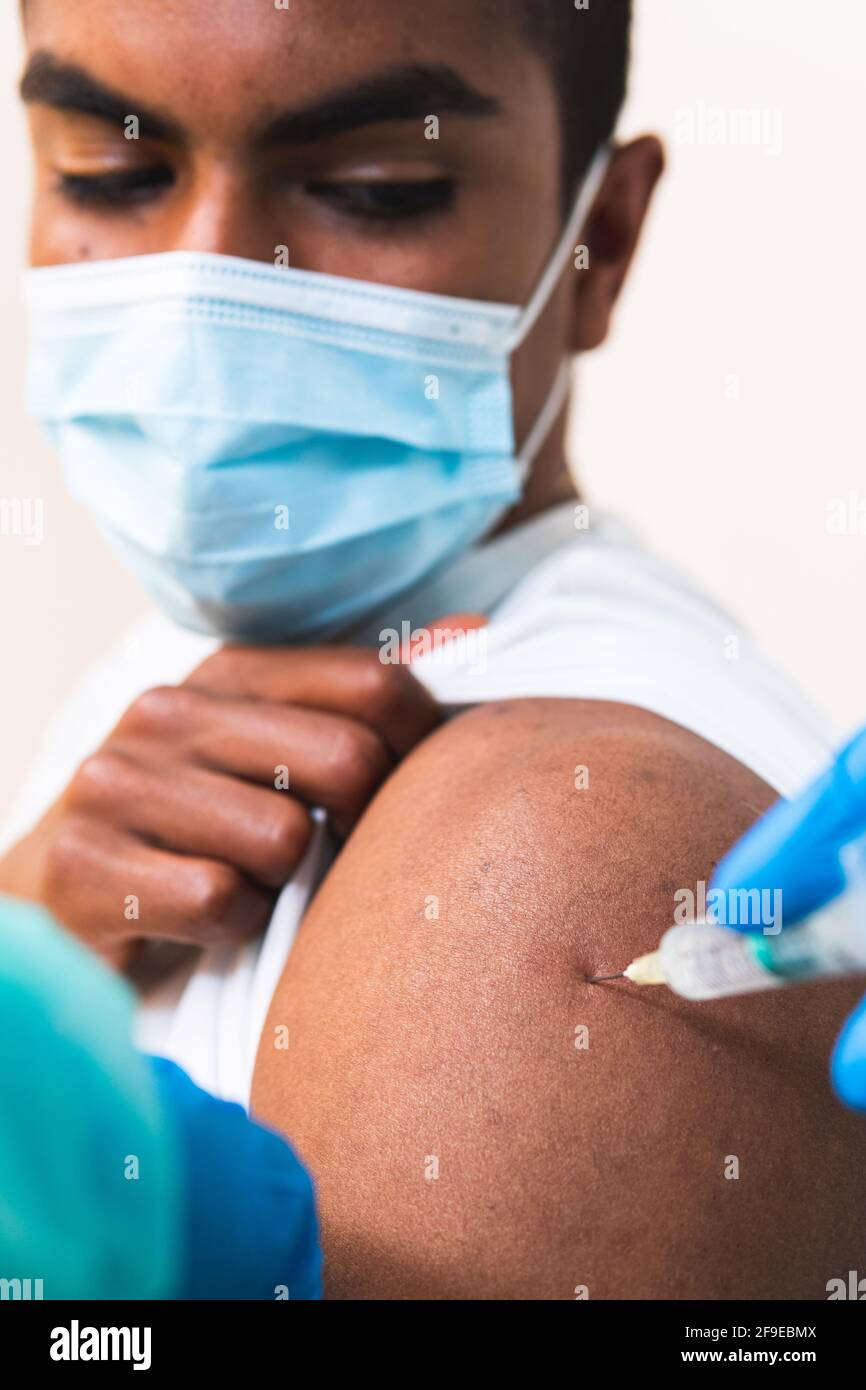 Spécialiste médical féminin en uniforme de protection, gants en latex et masque facial, non reconnaissable, en culture, qui vaccine un homme afro-américain en clinique Banque D'Images
