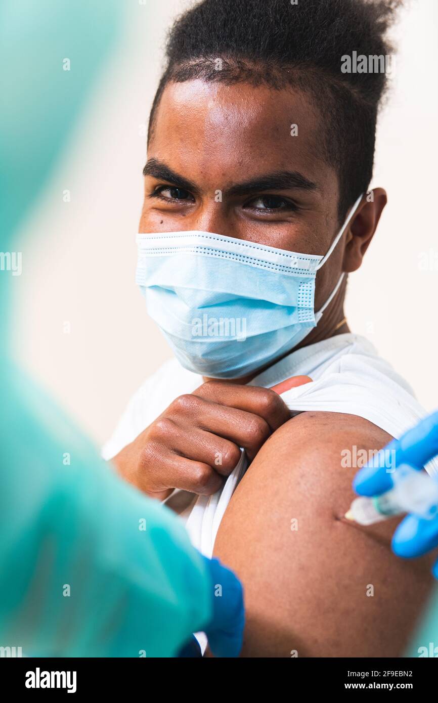 Spécialiste médical féminin en uniforme de protection, gants en latex et masque facial, non reconnaissable, en culture, qui vaccine un homme afro-américain en clinique Banque D'Images