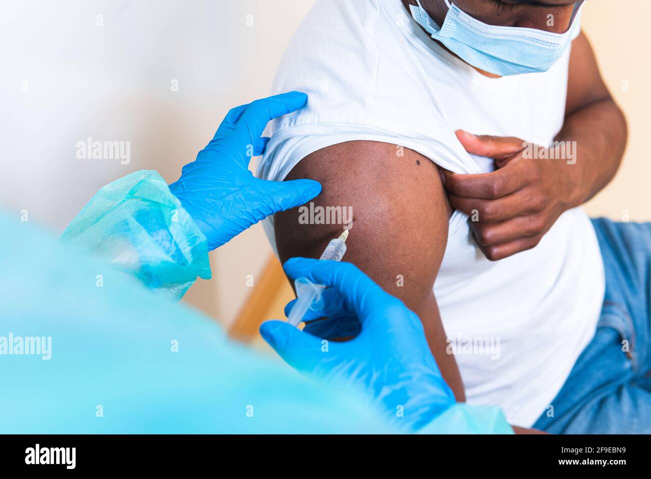 Spécialiste médical féminin en uniforme de protection, gants en latex et masque facial, non reconnaissable, en culture, qui vaccine un homme afro-américain en clinique Banque D'Images