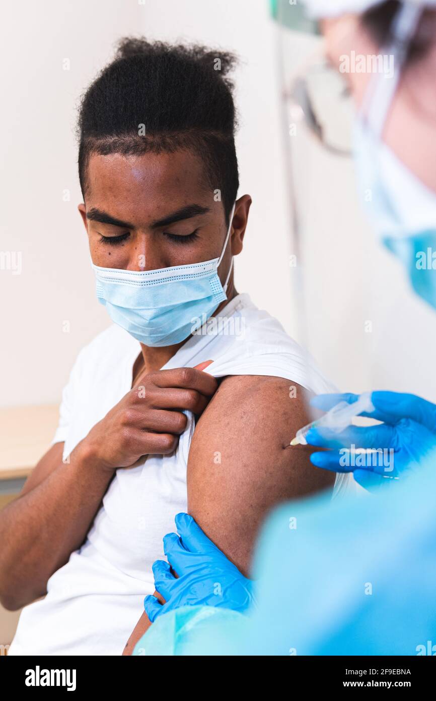 Spécialiste médical féminin en uniforme de protection, gants en latex et masque facial, non reconnaissable, en culture, qui vaccine un homme afro-américain en clinique Banque D'Images